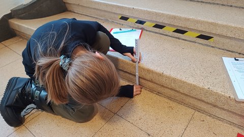 "Eine Person mit braunem Haar und grauem Haargummi sitzt auf einer Betontreppe und misst diese mit einem Stahllineal. Sie trägt dunkle Kleidung und schwarze Handschuhe. Auf der Treppenkante ist gelb-schwarzes Warnband angebracht. Ein Klemmbrett mit Papier und weitere Messinstrumente liegen neben der Person. Im Hintergrund Treppenstufen und eine Wand mit Fenster. Die Szene dokumentiert eine bauliche Überprüfung oder Messung."