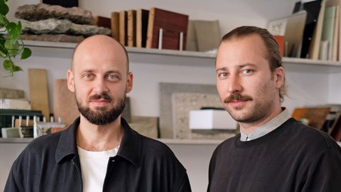 „Porträt zweier Männer, die nebeneinander stehen. Der linke Mann ist kahlköpfig mit Bart und trägt ein schwarzes Überhemd. Der rechte Mann hat zurückgebundene Haare, einen Schnäuzer und trägt einen schwarzen Pullover. Im Hintergrund sind Regale mit verschiedenen Materialmustern aus Stein und Holz zu sehen.“