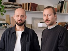 „Porträt zweier Männer, die nebeneinander stehen. Der linke Mann ist kahlköpfig mit Bart und trägt ein schwarzes Überhemd. Der rechte Mann hat zurückgebundene Haare, einen Schnäuzer und trägt einen schwarzen Pullover. Im Hintergrund sind Regale mit verschiedenen Materialmustern aus Stein und Holz zu sehen.“