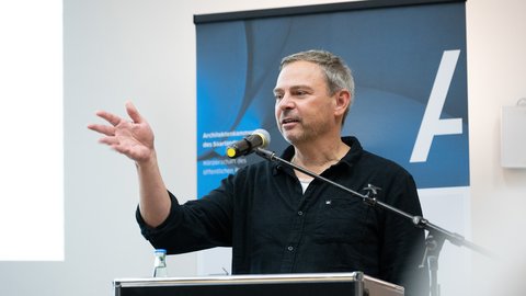 Eine einzelne Person steht an einem Rednerpult mit Mikrofon und gestikuliert während einer Ansprache, im Hintergrund ist ein Banner sichtbar, wodurch das Bild eine öffentliche Rede oder Veranstaltung dokumentiert und für fachlichen Austausch, Vermittlung von Positionen und aktive Beteiligung an architektonischem und gesellschaftlichem Diskurs steht.