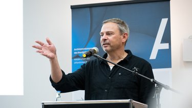 Eine einzelne Person steht an einem Rednerpult mit Mikrofon und gestikuliert während einer Ansprache, im Hintergrund ist ein Banner sichtbar, wodurch das Bild eine öffentliche Rede oder Veranstaltung dokumentiert und für fachlichen Austausch, Vermittlung von Positionen und aktive Beteiligung an architektonischem und gesellschaftlichem Diskurs steht.