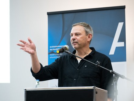Eine einzelne Person steht an einem Rednerpult mit Mikrofon und gestikuliert während einer Ansprache, im Hintergrund ist ein Banner sichtbar, wodurch das Bild eine öffentliche Rede oder Veranstaltung dokumentiert und für fachlichen Austausch, Vermittlung von Positionen und aktive Beteiligung an architektonischem und gesellschaftlichem Diskurs steht.