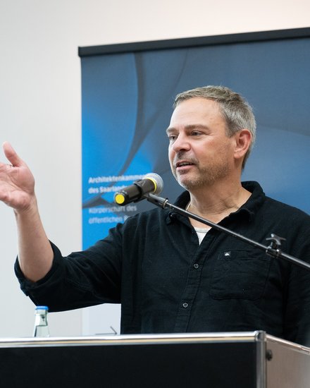 Eine einzelne Person steht an einem Rednerpult mit Mikrofon und gestikuliert während einer Ansprache, im Hintergrund ist ein Banner sichtbar, wodurch das Bild eine öffentliche Rede oder Veranstaltung dokumentiert und für fachlichen Austausch, Vermittlung von Positionen und aktive Beteiligung an architektonischem und gesellschaftlichem Diskurs steht.