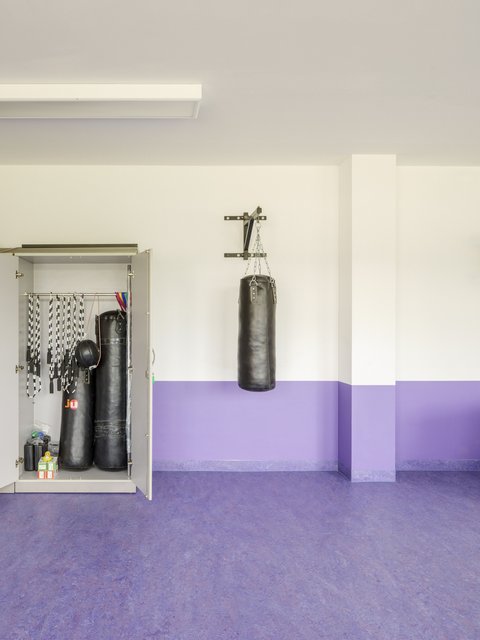 Eine Nahaufnahme im Sportraum. Links steht ein offener Metallschrank, in dem zwei schwarze Boxsäcke, Springseile und anderes Sportequipment lagern. Rechts davon hängt ein weiterer schwarzer Boxsack an einer Wandhalterung über dem lila Wandsockel.