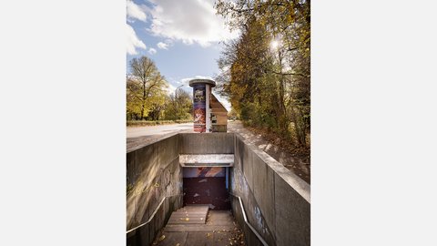 Nahansicht eines Unterführungsabgangs mit Geländern und Stufen; daneben Allee mit herbstlichem Laub, Litfaßsäule und kleines Holzhaus.