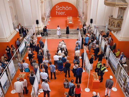 Luftaufnahme einer Ausstellung in barocker Kirche mit orange bestrachenem Boden. Ein großer oranger Bogen trägt weißen Text "Schön hier." Besucher unterschiedlichen Alters verteilen sich zwischen Ausstellungstafeln. Weiße Säulen und goldene Ornamente prägen den Raum. Ein Podium mit mehreren Personen ist zentral positioniert.