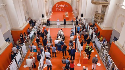 Luftaufnahme einer Ausstellung in barocker Kirche mit orange bestrachenem Boden. Ein großer oranger Bogen trägt weißen Text "Schön hier." Besucher unterschiedlichen Alters verteilen sich zwischen Ausstellungstafeln. Weiße Säulen und goldene Ornamente prägen den Raum. Ein Podium mit mehreren Personen ist zentral positioniert.