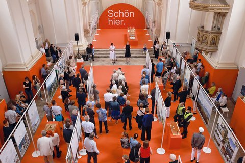 Luftaufnahme einer Ausstellung in barocker Kirche mit orange bestrachenem Boden. Ein großer oranger Bogen trägt weißen Text "Schön hier." Besucher unterschiedlichen Alters verteilen sich zwischen Ausstellungstafeln. Weiße Säulen und goldene Ornamente prägen den Raum. Ein Podium mit mehreren Personen ist zentral positioniert.