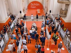 Luftaufnahme einer Ausstellung in barocker Kirche mit orange bestrachenem Boden. Ein großer oranger Bogen trägt weißen Text "Schön hier." Besucher unterschiedlichen Alters verteilen sich zwischen Ausstellungstafeln. Weiße Säulen und goldene Ornamente prägen den Raum. Ein Podium mit mehreren Personen ist zentral positioniert.