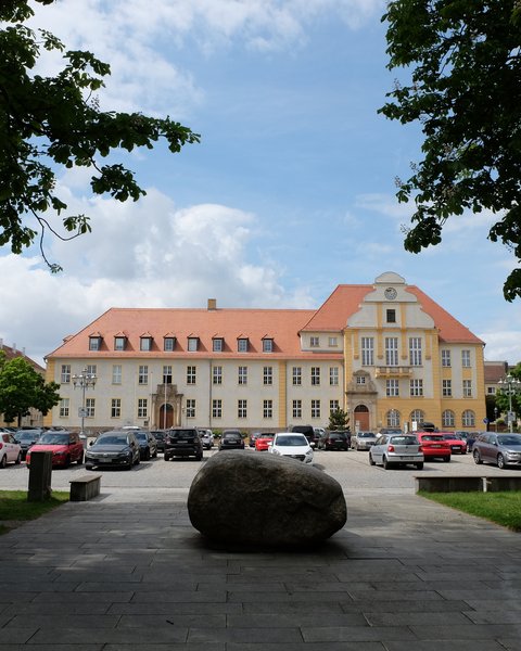 Großes historisches Rathausgebäude mit rotem Dach und gelber Fassade vor einem Parkplatz und einem großen Stein auf gepflastertem Weg im Vordergrund