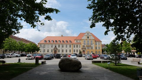 Großes historisches Rathausgebäude mit rotem Dach und gelber Fassade vor einem Parkplatz und einem großen Stein auf gepflastertem Weg im Vordergrund