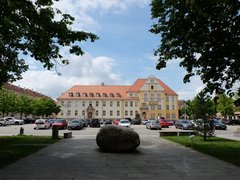 Großes historisches Rathausgebäude mit rotem Dach und gelber Fassade vor einem Parkplatz und einem großen Stein auf gepflastertem Weg im Vordergrund