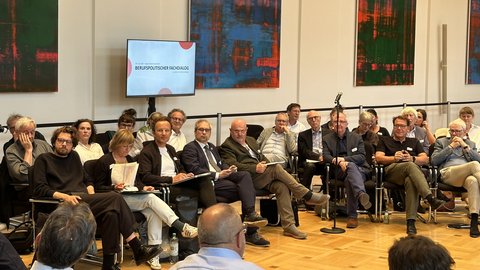 "Veranstaltungssaal mit Stuhlreihen, in denen etwa 20 Personen verschiedener Altersgruppen sitzen. Hinter ihnen an der Wand hängen vier abstrakte Gemälde mit Grün- und Blautönen sowie roten Akzenten. Im Raum ist ein Monitor sichtbar, auf dem der Text 'BERUFSPOLITISCHER FACHDIALOG' angezeigt wird. Ein Mikrofon befindet sich im Vordergrund. Der Raum hat Holzboden und moderne Wandgestaltung mit schwarzem Handlauf."
