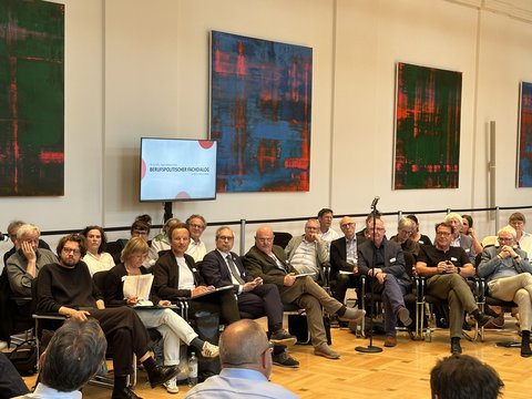 "Veranstaltungssaal mit Stuhlreihen, in denen etwa 20 Personen verschiedener Altersgruppen sitzen. Hinter ihnen an der Wand hängen vier abstrakte Gemälde mit Grün- und Blautönen sowie roten Akzenten. Im Raum ist ein Monitor sichtbar, auf dem der Text 'BERUFSPOLITISCHER FACHDIALOG' angezeigt wird. Ein Mikrofon befindet sich im Vordergrund. Der Raum hat Holzboden und moderne Wandgestaltung mit schwarzem Handlauf."