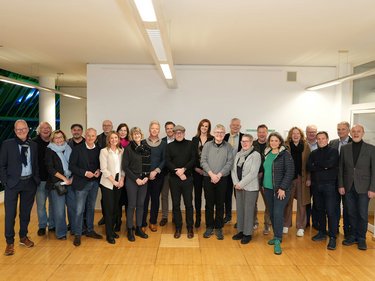 Eine größere Gruppe von etwa 25 bis 30 erwachsenen Personen steht dicht nebeneinander im selben Raum, locker angeordnet und unterschiedlich gekleidet, viele lächeln oder wirken entspannt, wodurch das Foto eine gemeinschaftliche Situation dokumentiert und für kollektive Zusammenarbeit, fachlichen Austausch und die Vielfalt beteiligter Akteurinnen und Akteure innerhalb eines gemeinsamen Projekts oder Netzwerks steht.