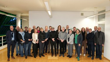Eine größere Gruppe von etwa 25 bis 30 erwachsenen Personen steht dicht nebeneinander im selben Raum, locker angeordnet und unterschiedlich gekleidet, viele lächeln oder wirken entspannt, wodurch das Foto eine gemeinschaftliche Situation dokumentiert und für kollektive Zusammenarbeit, fachlichen Austausch und die Vielfalt beteiligter Akteurinnen und Akteure innerhalb eines gemeinsamen Projekts oder Netzwerks steht.
