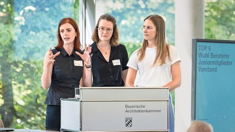 „Drei Frauen präsentieren gemeinsam am Rednerpult der Bayerischen Architektenkammer. Auf einem Bildschirm steht: ‚Top 6 Wahl Beraterin Juniormitglieder Vorstand‘.“