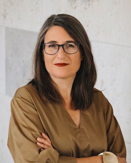 Porträtfoto einer Frau mit schwarzen, schulterlanken Haaren und Brille. Sie trägt ein beiges Leinenhemd und hat ihre Arme verschränkt. Sie blickt direkt in die Kamera und hat rote Lippen. Der Hintergrund zeigt eine helle Wand mit geometrischen, hellen Fliesenmuster.