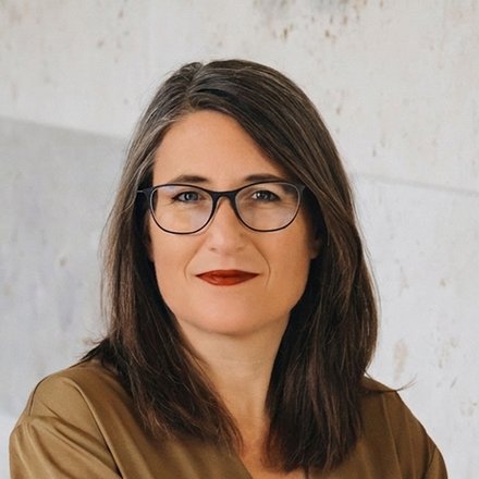 Porträtfoto einer Frau mit schwarzen, schulterlanken Haaren und Brille. Sie trägt ein beiges Leinenhemd und hat ihre Arme verschränkt. Sie blickt direkt in die Kamera und hat rote Lippen. Der Hintergrund zeigt eine helle Wand mit geometrischen, hellen Fliesenmuster.