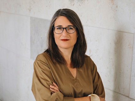Porträtfoto einer Frau mit schwarzen, schulterlanken Haaren und Brille. Sie trägt ein beiges Leinenhemd und hat ihre Arme verschränkt. Sie blickt direkt in die Kamera und hat rote Lippen. Der Hintergrund zeigt eine helle Wand mit geometrischen, hellen Fliesenmuster.