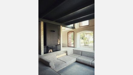 Wohnbereich: Niedrige Ecksofa‑Landschaft vor einem schlichten Kamin; große Rundbogenöffnungen zum Garten; Sichtbeton und Stahlträger.