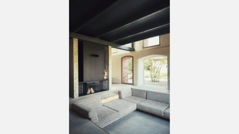 Wohnbereich: Niedrige Ecksofa‑Landschaft vor einem schlichten Kamin; große Rundbogenöffnungen zum Garten; Sichtbeton und Stahlträger.