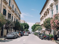 Eine breite Straße in südlichem Stil mit Klassizismus-Gebäuden beidseitig. Mehrere Autos sind geparkt, Motorräder stehen rechts. Blühende Bäume mit rosa und weißen Blüten säumen beide Seiten. Ein klarer, blauer Himmel mit wenigen Wolken.
