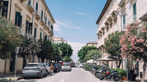 Eine breite Straße in südlichem Stil mit Klassizismus-Gebäuden beidseitig. Mehrere Autos sind geparkt, Motorräder stehen rechts. Blühende Bäume mit rosa und weißen Blüten säumen beide Seiten. Ein klarer, blauer Himmel mit wenigen Wolken.