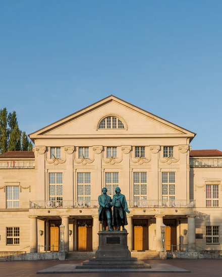 Schloss in Beige mit vielen Fenstern und blauem Himmel und einem Baum der hinter dem linken Flügel hervorschaut 