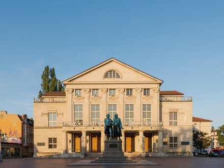 Schloss in Beige mit vielen Fenstern und blauem Himmel und einem Baum der hinter dem linken Flügel hervorschaut 
