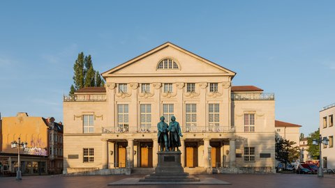 Schloss in Beige mit vielen Fenstern und blauem Himmel und einem Baum der hinter dem linken Flügel hervorschaut 