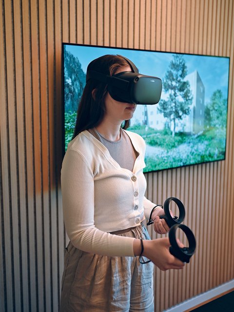 "Eine junge Frau mit langen braunen Haaren trägt eine schwarze Virtual-Reality-Brille und hält schwarze VR-Controller in beiden Händen. Sie trägt einen cremefarbenen Strickpullover mit Knopfleiste über einem hellgrauen T-Shirt und eine hellbeige Leinenhose. Sie blickt nach oben, während sie die VR-Erfahrung nutzt. Hinter ihr zeigt ein großer Bildschirm an der Wand eine Architekturvisualisierung mit grünen Wiesen, Bäumen und einem modernen Gebäude unter blauem Himmel. Die Wand dahinter ist mit vertikalen Holzlamellen in warmen Natur- und Grautönen verkleidet."