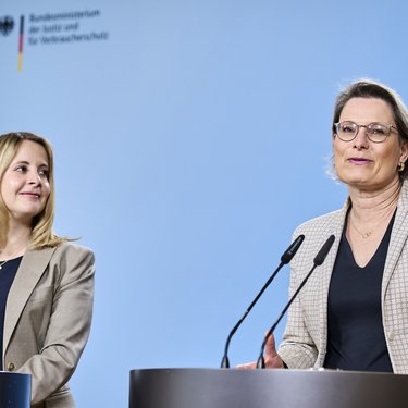 Zwei Frauen stehen an Redepulten vor hellblauem Hintergrund und sprechen in Mikrofone; eine Frau blickt zur Seite, die andere spricht direkt ins Publikum, im Hintergrund das Logo eines Bundesministeriums.
