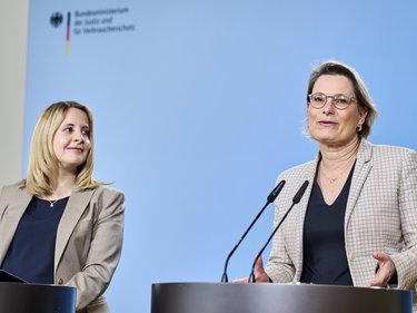 Zwei Frauen stehen an Redepulten vor hellblauem Hintergrund und sprechen in Mikrofone; eine Frau blickt zur Seite, die andere spricht direkt ins Publikum, im Hintergrund das Logo eines Bundesministeriums.