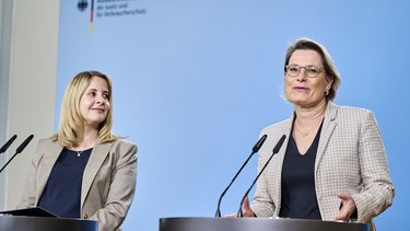 Zwei Frauen stehen an Redepulten vor hellblauem Hintergrund und sprechen in Mikrofone; eine Frau blickt zur Seite, die andere spricht direkt ins Publikum, im Hintergrund das Logo eines Bundesministeriums.