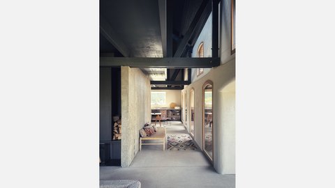 Langer Flur im Feldsteinhaus: Sichtbetonwände und dunkelgraue Stahlträger, rechts bogenförmige Holzfenster, hinten offene Küche; Bank und bunter Teppichläufer.