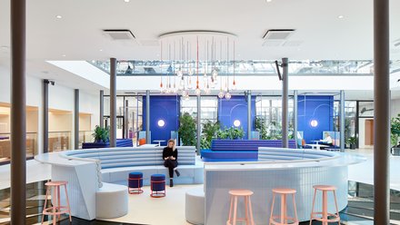 Offener Innenraum mit Sitzlandschaften: Runde, gepolsterte Sitzmöbel in Blau- und Pastelltönen, Pflanzen und hängende Leuchten in hellem Atrium.