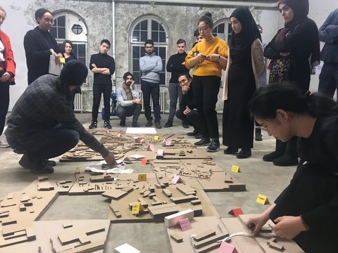 Eine Gruppe steht und hockt um ein großes Stadtmodell aus Pappe. Zwei Teilnehmende bearbeiten das Modell, während die anderen die markierten Gebäude und Flächen betrachten.