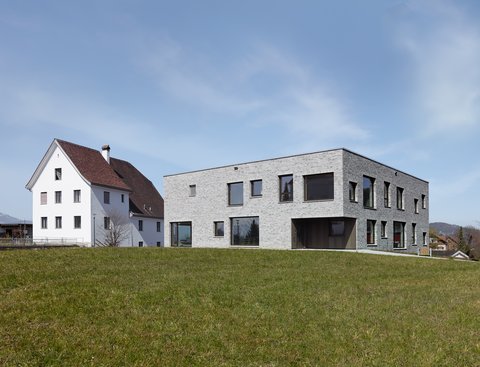Modernes zweistöckiges Gebäude mit grauer Steinfassade neben einem traditionellen weißen Haus mit rotem Dach auf einer grünen Wiese