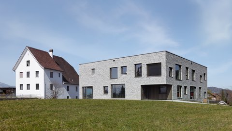 Modernes zweistöckiges Gebäude mit grauer Steinfassade neben einem traditionellen weißen Haus mit rotem Dach auf einer grünen Wiese