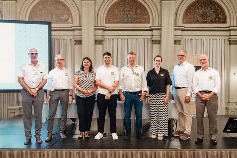 "Zehn Personen stehen nebeneinander auf einer Bühne vor klassischer Architektur mit Säulen und goldenen Verzierungen. Hinter der Gruppe ist eine Präsentationsfläche sichtbar. Die meisten tragen weiße oder helle Oberteile, viele mit bunten Anstecknadeln. Eine Frau trägt ein Muster-Shirt. Formale Veranstaltungsatmosphäre."