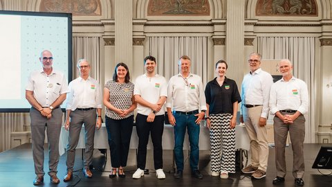 "Zehn Personen stehen nebeneinander auf einer Bühne vor klassischer Architektur mit Säulen und goldenen Verzierungen. Hinter der Gruppe ist eine Präsentationsfläche sichtbar. Die meisten tragen weiße oder helle Oberteile, viele mit bunten Anstecknadeln. Eine Frau trägt ein Muster-Shirt. Formale Veranstaltungsatmosphäre."