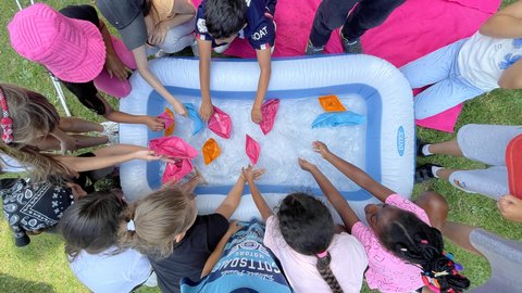 "Blick von oben auf eine Gruppe von Kindern, die im Gras um ein kleines aufblasbares Wasserbecken stehen. Mit den Händen setzen sie bunte Papierschiffchen in das Wasser."