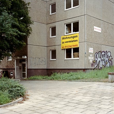 Foto Plattenbau mit Banner Alt: Hausecke eines grauen Plattenbaus; gelbes Schild „Wohnungen zu vermieten, tel. 015256270493“ am Fenster; Graffiti; Eingang links.