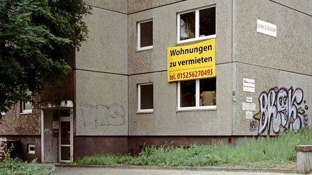Foto Plattenbau mit Banner Alt: Hausecke eines grauen Plattenbaus; gelbes Schild „Wohnungen zu vermieten, tel. 015256270493“ am Fenster; Graffiti; Eingang links.