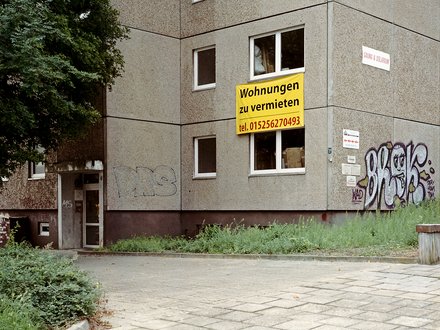 Foto Plattenbau mit Banner Alt: Hausecke eines grauen Plattenbaus; gelbes Schild „Wohnungen zu vermieten, tel. 015256270493“ am Fenster; Graffiti; Eingang links.