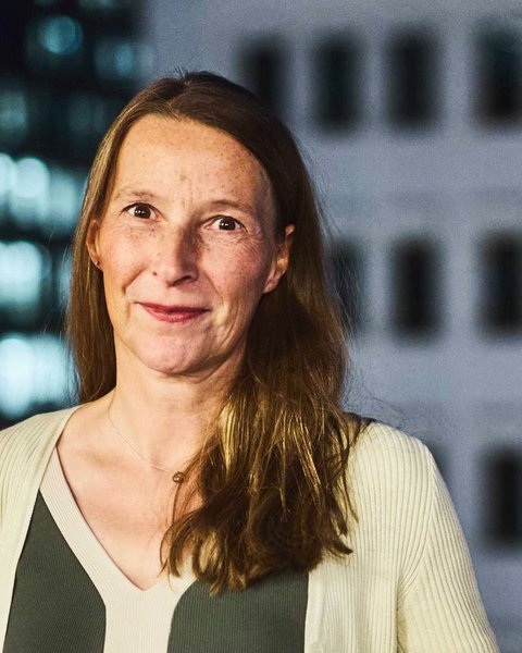 "Porträt einer Frau mit mittellangen, braunen Haaren und Sommersprossen. Sie trägt ein dunkelgrünes Oberteil unter einer hellen, gerippten Strickjacke und blickt lächelnd in die Kamera. Im Hintergrund ist ein unscharfes Hochhaus mit beleuchteten Fenstern und Fassadenelementen bei Nacht zu sehen."