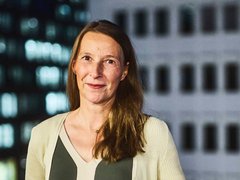 "Porträt einer Frau mit mittellangen, braunen Haaren und Sommersprossen. Sie trägt ein dunkelgrünes Oberteil unter einer hellen, gerippten Strickjacke und blickt lächelnd in die Kamera. Im Hintergrund ist ein unscharfes Hochhaus mit beleuchteten Fenstern und Fassadenelementen bei Nacht zu sehen."