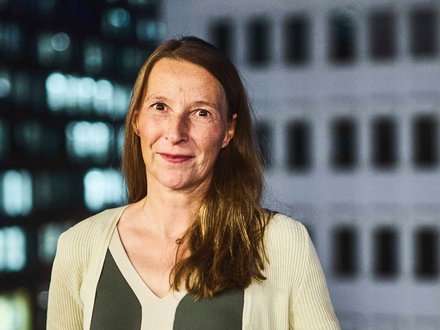 "Porträt einer Frau mit mittellangen, braunen Haaren und Sommersprossen. Sie trägt ein dunkelgrünes Oberteil unter einer hellen, gerippten Strickjacke und blickt lächelnd in die Kamera. Im Hintergrund ist ein unscharfes Hochhaus mit beleuchteten Fenstern und Fassadenelementen bei Nacht zu sehen."