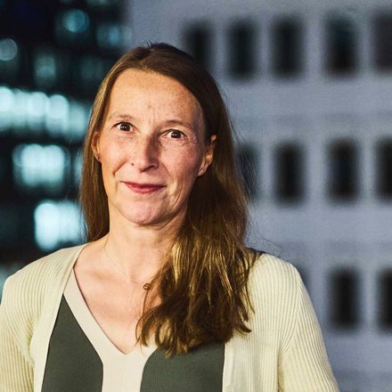 "Porträt einer Frau mit mittellangen, braunen Haaren und Sommersprossen. Sie trägt ein dunkelgrünes Oberteil unter einer hellen, gerippten Strickjacke und blickt lächelnd in die Kamera. Im Hintergrund ist ein unscharfes Hochhaus mit beleuchteten Fenstern und Fassadenelementen bei Nacht zu sehen."